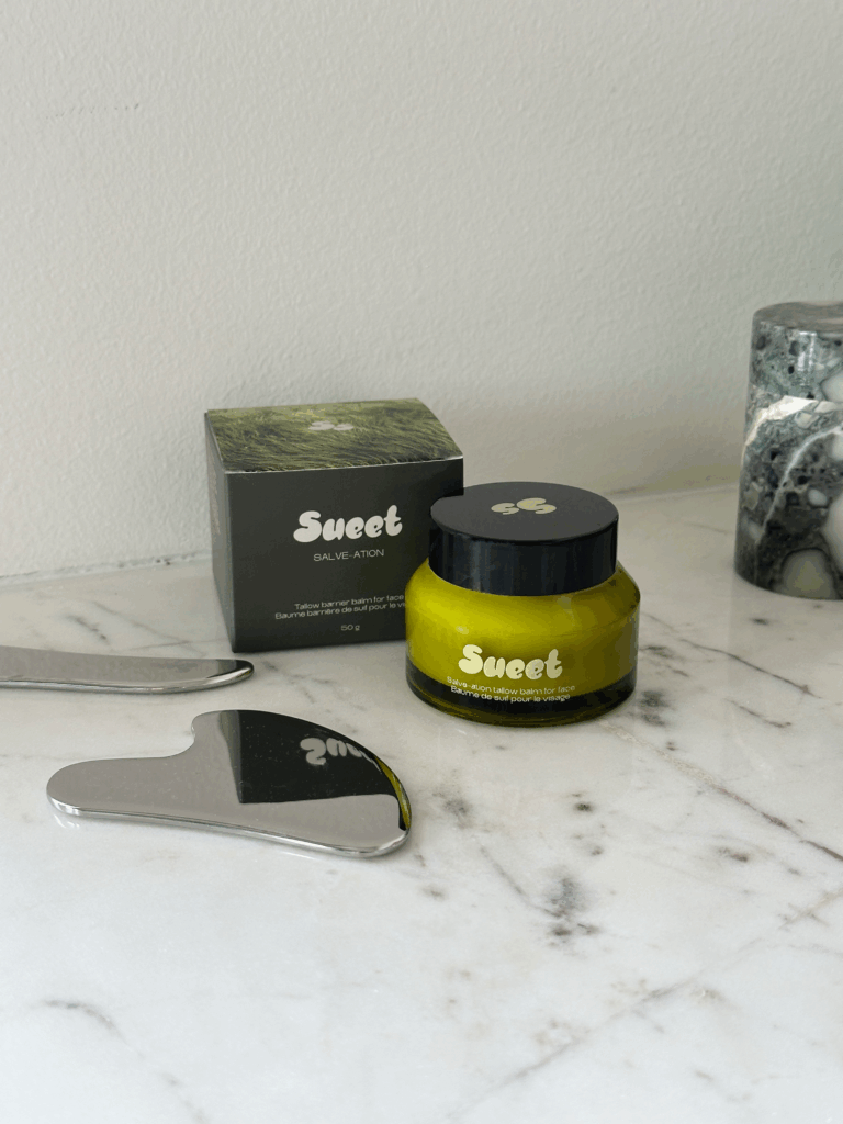 Sueet Skin Salve-ation Tallow Balm on a marble counter surrounded by the brand's stainless steel gua-sha.