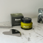 Sueet Skin Salve-ation Tallow Balm on a marble counter surrounded by the brand's stainless steel gua-sha.