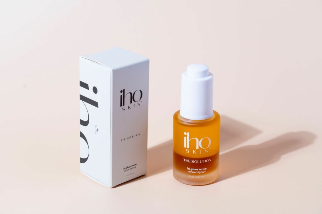 A bottle of Iho's serum next to it's white and black packaging, in front of a light pink background