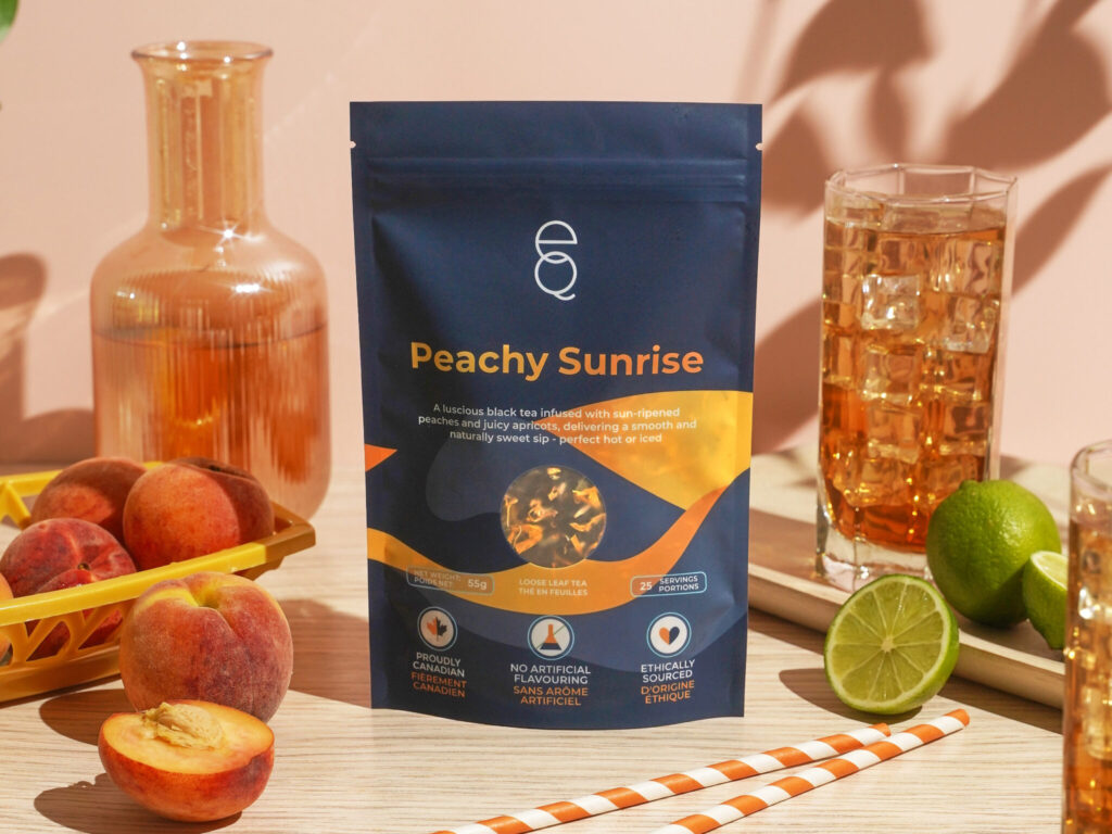 A blue bag of "Peachy Sunrise" tea sitting on a counter surrounded by a basket of peaches, some limes, some brewed iced tea, and a set of orange and white striped straws.