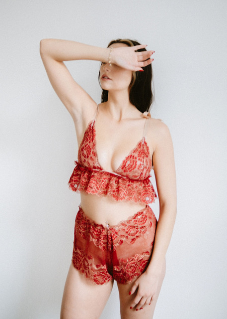 A model standing with her arm covering her face, wearing a bra and high-waisted boxers set in a sheer red lace.