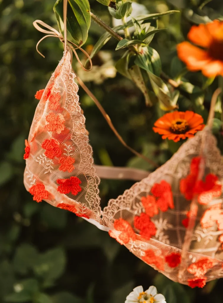 A sheer cream coloured bralette with vibrant orange-red poppies embroidered on it, handing from a brand with red poppies in the background.